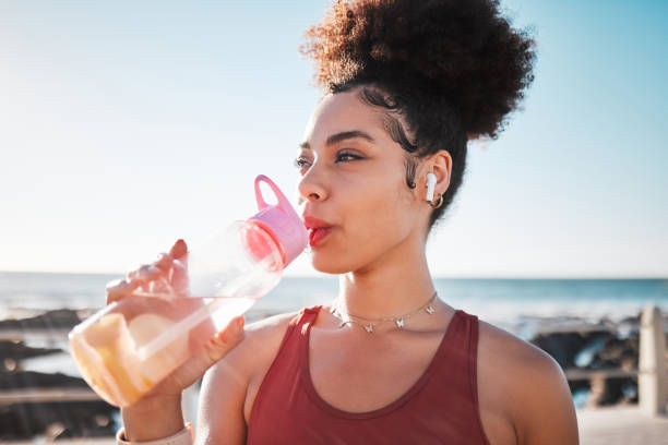 運動のためにビーチで水を飲み、音楽を聴き、青い空のレンズフレアで有酸素運動をするフィットネス黒人女性。 ダイエット、目標、疲れたスポーツランナー、または海でのアメリカの人の - exercising sports training lens flare women ストックフォトと画像