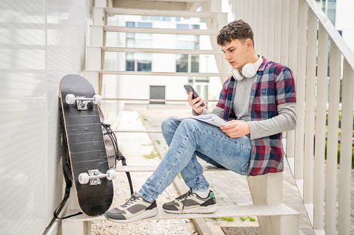 One Young man teenager caucasian student with books read study at campus in day sit on staircase education concept copy space use smartphone mobile phone for online text or sms app social network