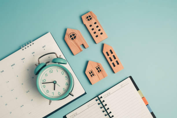 un reloj despertador con calendario, cuaderno y modelo de casa de madera - currency house banking real estate fotografías e imágenes de stock