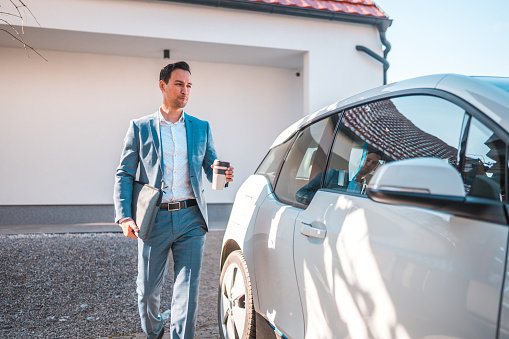 Morning outside of a modern home. Asian mature man in a blue suit walking to his electric car to get to work on time.