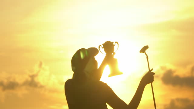 Golf winner holding up golden trophy cup in hand at sunset silhouette.