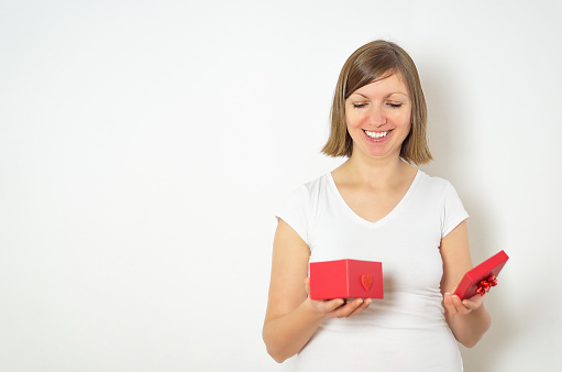 Portrait of a happy smiling woman in a white shirt opening a red present box for the Valentines day. High quality photo.