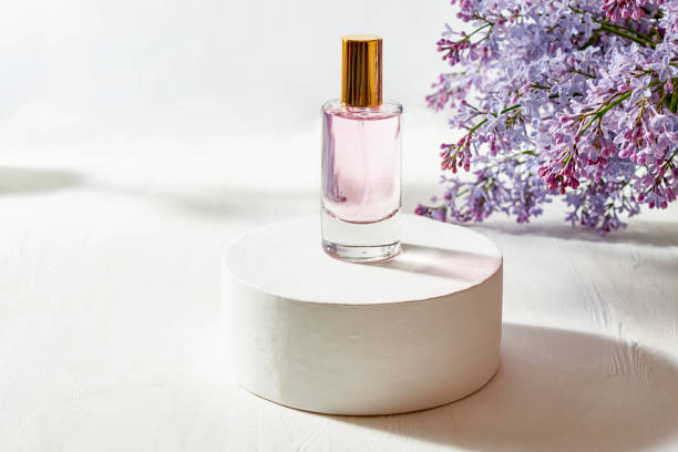 istock Pink glass perfume bottle on white round podium and lilac branches 1484595080