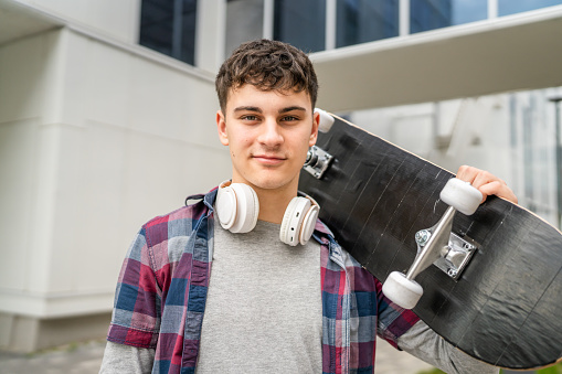 One man young adult caucasian teenager stand outdoor with skateboard on his shoulder and headphones posing portrait looking to the camera happy confident wear shirt casual real person copy space