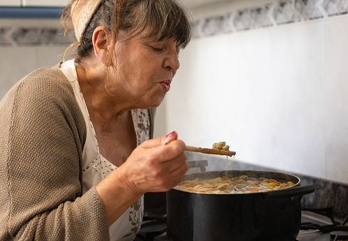 mature senior woman cooking on the kitchen stove. typical spanish dish in a pot. beans with chorizo, potatoes, vegetables and iberian sausage. blow because it is hot.