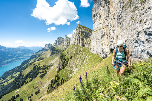 Mountaineer hiking on flowery meadow below steep rock wall with scenic view on lake Walensee and the Churfürsten mountain range. Schnürliweg, Walensee, St. Gallen, Switzerland, Europe.