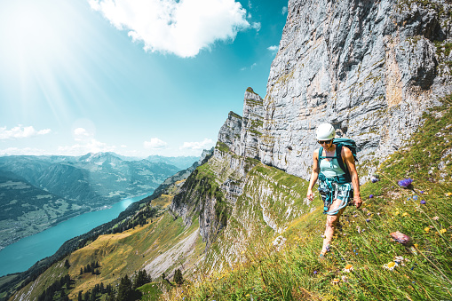 Atheltic woman hiking on flowery trail next to steep rock wall with scenic view on Walensee and Churfürsten mountain range in the background. Schnürliweg, Walensee, St. Gallen, Switzerland, Europe.