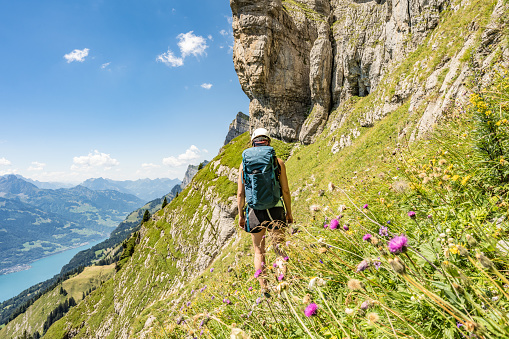 Back view of sportive woman hiking on flowery trail next to steep rock wall with scenic view on Walensee and the swiss alps in the background. Schnürliweg, Walensee, St. Gallen, Switzerland, Europe.