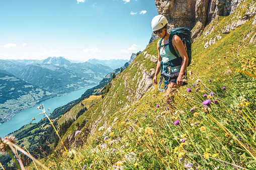 Side view of sportive woman hiking on flowery trail next to steep rock wall with scenic view on Walensee and the swiss alps in the background. Schnürliweg, Walensee, St. Gallen, Switzerland, Europe.