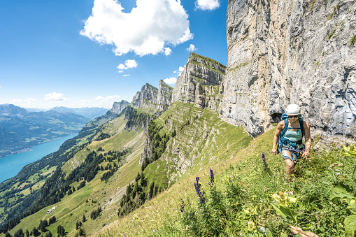 Athletic woman hiking on flowery meadow below steep rock wall with scenic view on lake Walensee and the swiss alps. Schnürliweg, Walensee, St. Gallen, Switzerland, Europe.