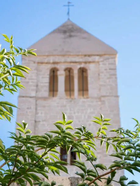 Rab, Croatia - August 24, 2022: A stunning bell tower of a church in Rab, Croatia stands out against the blue sky. Leaf trees and plants line its walls as proof of belief. One of the five churches on Rab.