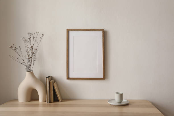 istock Empty wooden picture frame mockup hanging on beige wall background. Boho shaped vase, dry flowers on table. Cup of coffee, old books. Working space, home office. Art, poster display. Modern interior. 1484653629