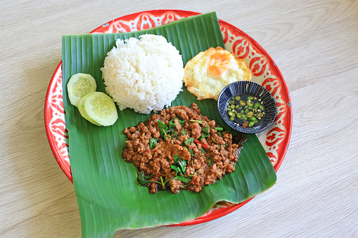 Stir-fried minced pork with basil leaves served with rice and fried egg on banana leaf on zinc tray over wooden table background.