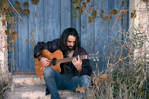 young caucasian man with long hair sitting in the doorway of a house concentrated playing the guitar - Macho lifestyle