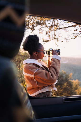 young african woman taking photos of nature with a camera during a road trip in camper van, concept of van life and travel adventures
