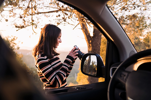 smiling caucasian woman taking photos of nature with a camera during a road trip in camper van, concept of van life and travel adventure