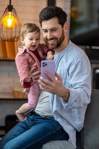 Happy dad and toddler child looking at cellphone, father holding phone taking selfie with cute little girl kid at home.