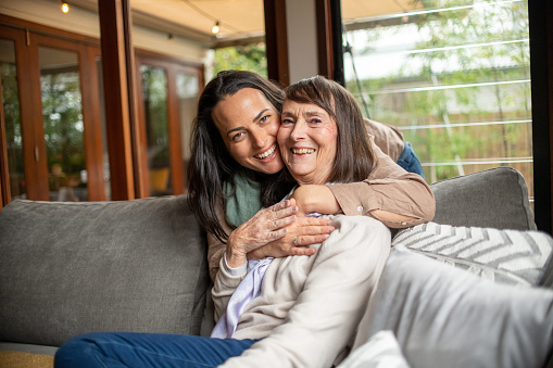 An attractive daughter warmly embraces her mother from behind. Both woman are smiling and looking directly at the camera. It's mother's day and they have spent the afternoon together at home.