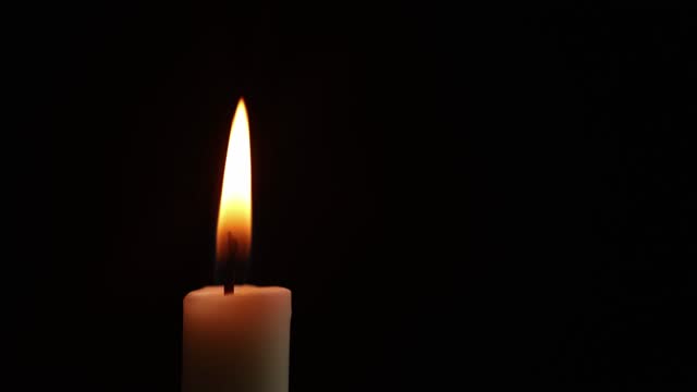 Close-up frame. A single wax white candle is burning. Orange light flame. The wind blows back and forth. black darkness background night