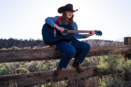 Woman Playing Guitar Sitting on Fence - Cowgirl wearing western attire sitting on wooden rural fence on ranch playing guitar.