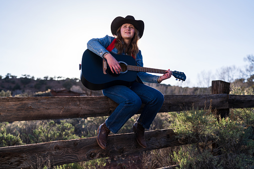 Woman Playing Guitar Sitting on Fence - Cowgirl wearing western attire sitting on wooden rural fence on ranch playing guitar.