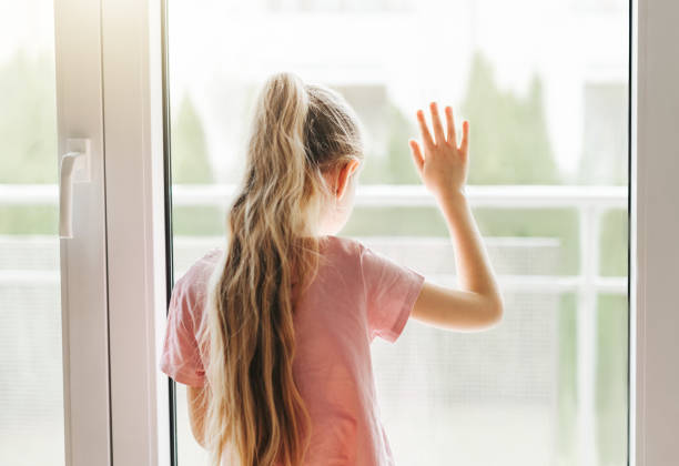 istock Sad little girl looking through  window at home. 1484719215