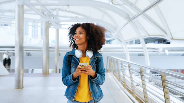 счастливая молодая афро-женщина слушает музыку плейлиста с беспроводными наушниками в желтой рубашке и джинсовой куртке на открытом возду - life jacket audio стоковые фото и изображения