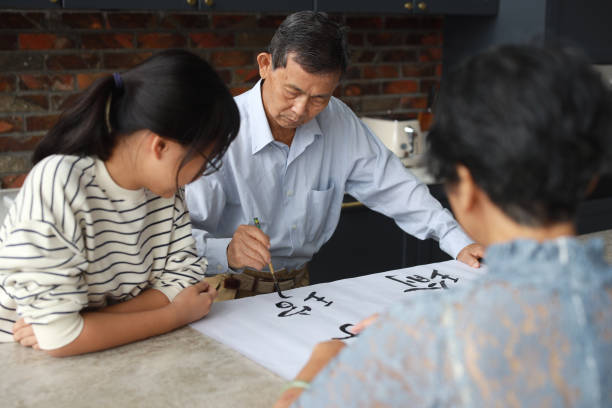 an asian grandfather is seen teaching and showing his calligraphy skills to his wife and granddaughter at home. the atmosphere is filled with warmth and love as the family members engage in this traditional chinese activity together. - chinese script text calligraphy grandmother imagens e fotografias de stock