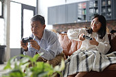 A cheerful Asian grandfather and his granddaughter are sitting on the sofa in the living room, having fun playing video games together at home. This heartwarming scene embodies the happy retirement concept