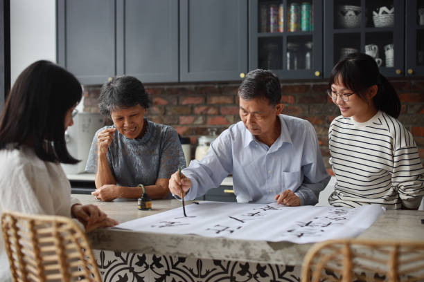 an asian grandfather is seen teaching and showing his calligraphy skills to his wife and two granddaughters at home. the atmosphere is filled with warmth and love as the family members engage in this traditional chinese activity together. - chinese script text calligraphy grandmother imagens e fotografias de stock