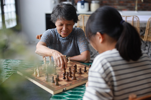 A cheerful Asian grandmother and her granddaughter are playing a game of chess together at home. This heartwarming scene embodies the concepts of happy retirement and bonding between generations