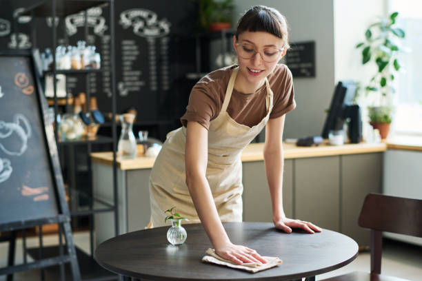 istock Young smiling waitress in apron bending over table while cleaning it with wipe 1484742234