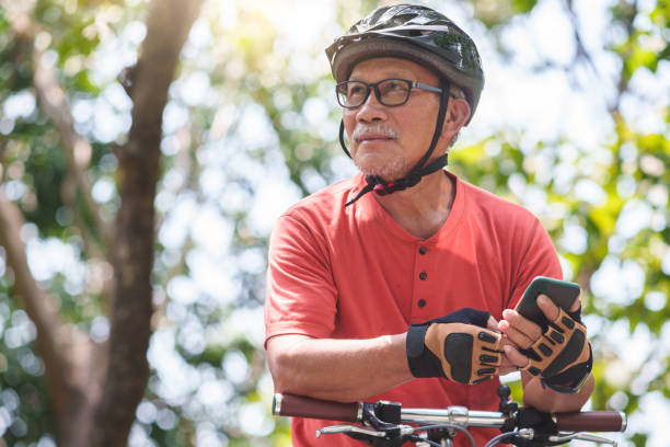 homme âgé asiatique tenant un téléphone portable - cycling senior adult sports helmet men photos et images de collection