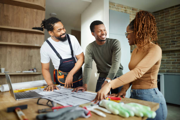 Happy black couple and manual worker working on blueprints at home. Happy African American couple talking while analyzing blueprints with manual worker in the apartment. renovation stock pictures, royalty-free photos & images