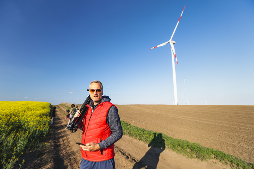 Man standing in the wind farm agricultural field with smart phone in hand, carrying tripod stand on shoulder, photographer freelancer