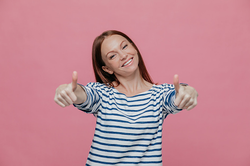 Joyful beautiful young lady raises thumbs at camera, shows that everything is okay, wears striped clothes, tilts head, smiles broadly, shows white teeth, isolated over pink background, says yes