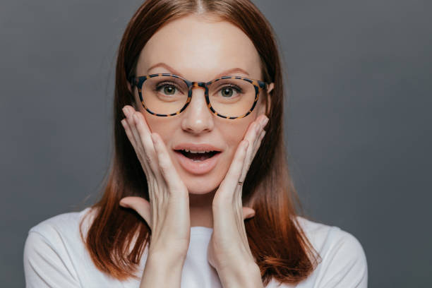 istock Headshot of surprised young Caucasian lady keeps both palms on her cheeks, looks with amazement, recieves unexpected news, wears spectacles, white clothing, poses alone against grey background 1484759901