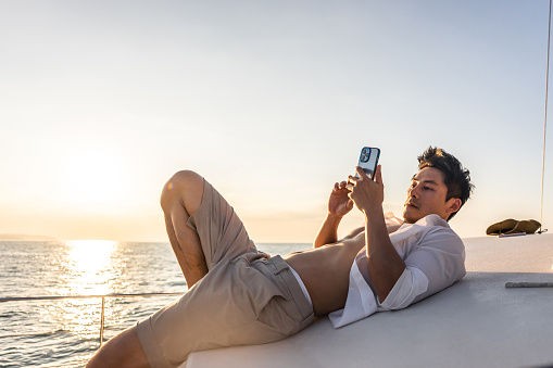 Asian young man tourist using smartphone taking photo during yachting. Attactive male feel happy and relax while sitting alone on deck of yacht, enjoy time when catamaran boat sailing during summer.