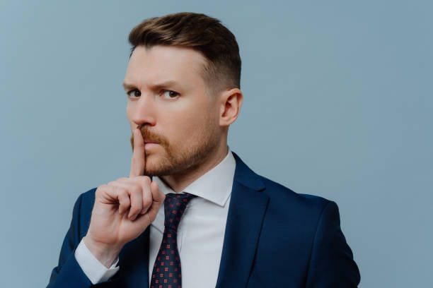istock Serious bearded prosperous male office worker dressed in formal clothes makes hush gesture asks to keep quiet demands silence with strict expression isolated over blue background. Taboo shh shushing 1484763804