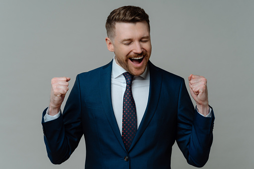 Cheerful excited bearded businessman makes yes gesture clenches fists feels like triumphant wears formal suit with tie celebrates success isolated over grey background. Yeah finally success.