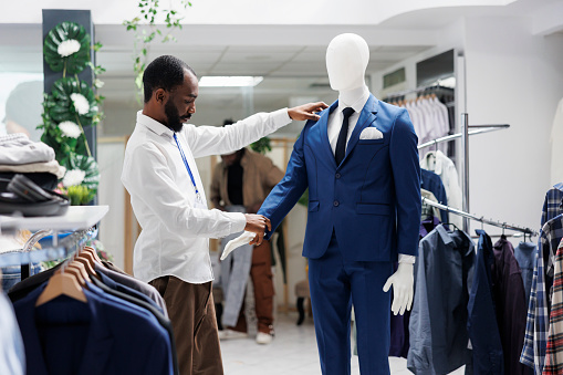 Boutique assistant checking mannequin dressed in formal menswear latest collection. African american man examining male luxury suit on model, while working in shopping mall