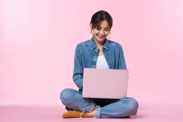 istock Young Asian college student sits cross-legged on floor working with a laptop computer, looking at the laptop monitor in a studio shot on a pink background 1484776946