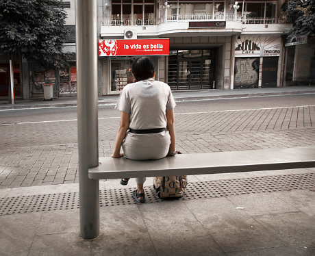 Madrid,Spain - October 4, 2011: rear view of one woman sitting alone at the bus stop, city street, centre of Madrid. Life is beutiful banner in spanish language in the background