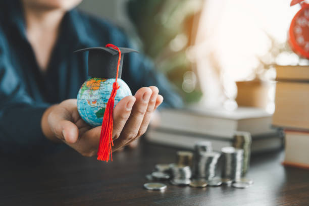 istock Hand holding graduation cap on globe with stack coin. Concept of saving money for a scholarship to study abroad at a university level. Financial planning accounting ideas for future education. 1484781461