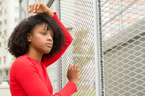 Serious and introspective young Afro woman leaning on a wire fence, lost in thought and contemplation