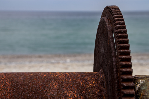 Old rusty cogwheel wich was a part of a winch. Sea and beach on background. 
Сoncept of sea influence on steel mechanisms