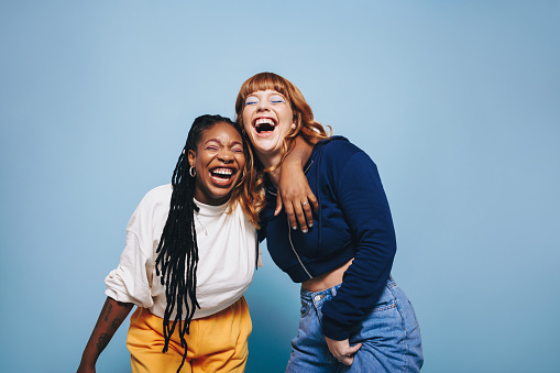 Interracial best friends laughing and having a good time together in a studio. Happy young women enjoying themselves while standing against a blue background. Two vibrant female friends making memories.
