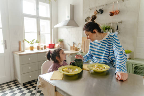 It's lunch time! Photo of a little girl eating pasta for lunch while sitting in high chair with her mom, in their family kitchen kids eating noodles stock pictures, royalty-free photos & images