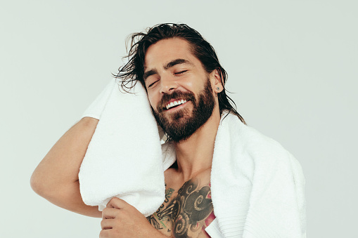 Refreshed young man wiping his hair with a bath towel after taking a relaxing shower. With a smile on his face, a handsome caucasian man enjoys his grooming and self-care routine in a studio.