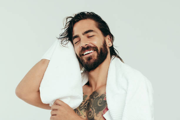 Refreshed young man using a bath towel after a shower Refreshed young man wiping his hair with a bath towel after taking a relaxing shower. With a smile on his face, a handsome caucasian man enjoys his grooming and self-care routine in a studio. body care shower stock pictures, royalty-free photos & images